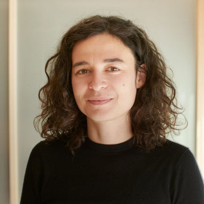 Porträt einer jungen Frau mit braunen, lockigen Haaren und dezentem Lächeln, schwarzes Oberteil, unscharfer Hintergrund.