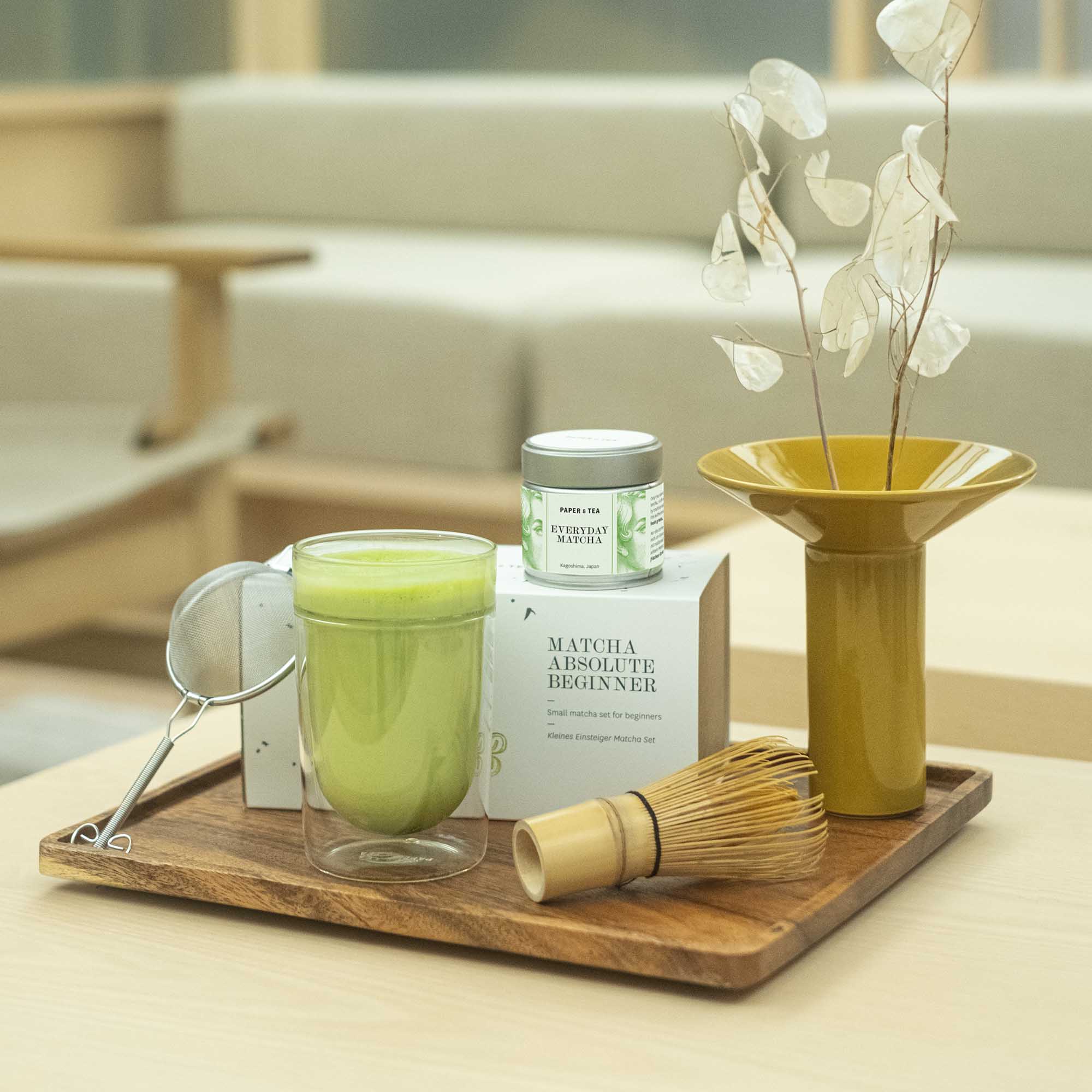 Holztablett mit Matcha-Einsteiger-Set, grünem Matcha im Glas und gelber Keramikvase mit getrockneten Blättern.
