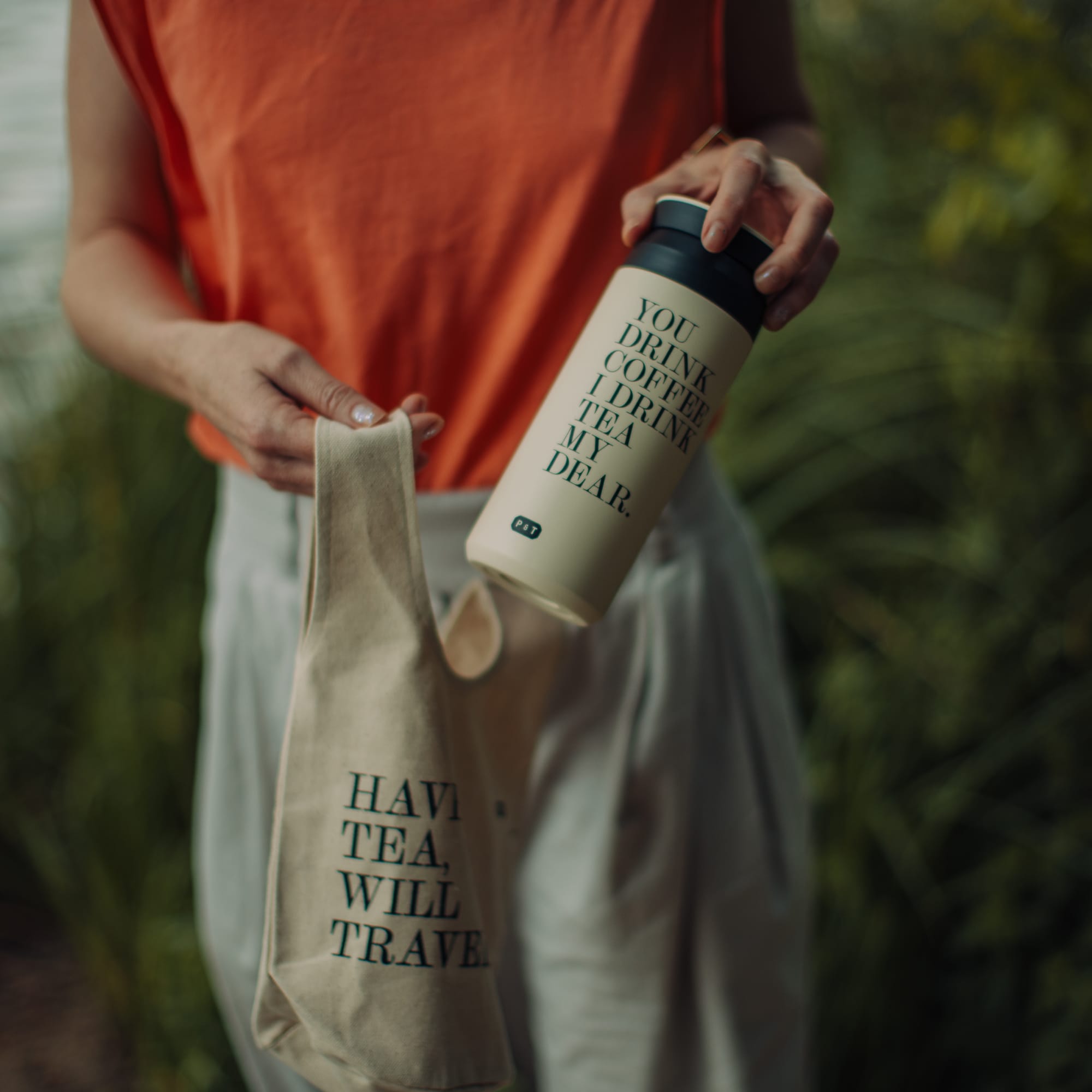 Person im orangefarbenen Top hält cremefarbenen Isolierbecher mit Aufschrift vor grüner Pflanze.