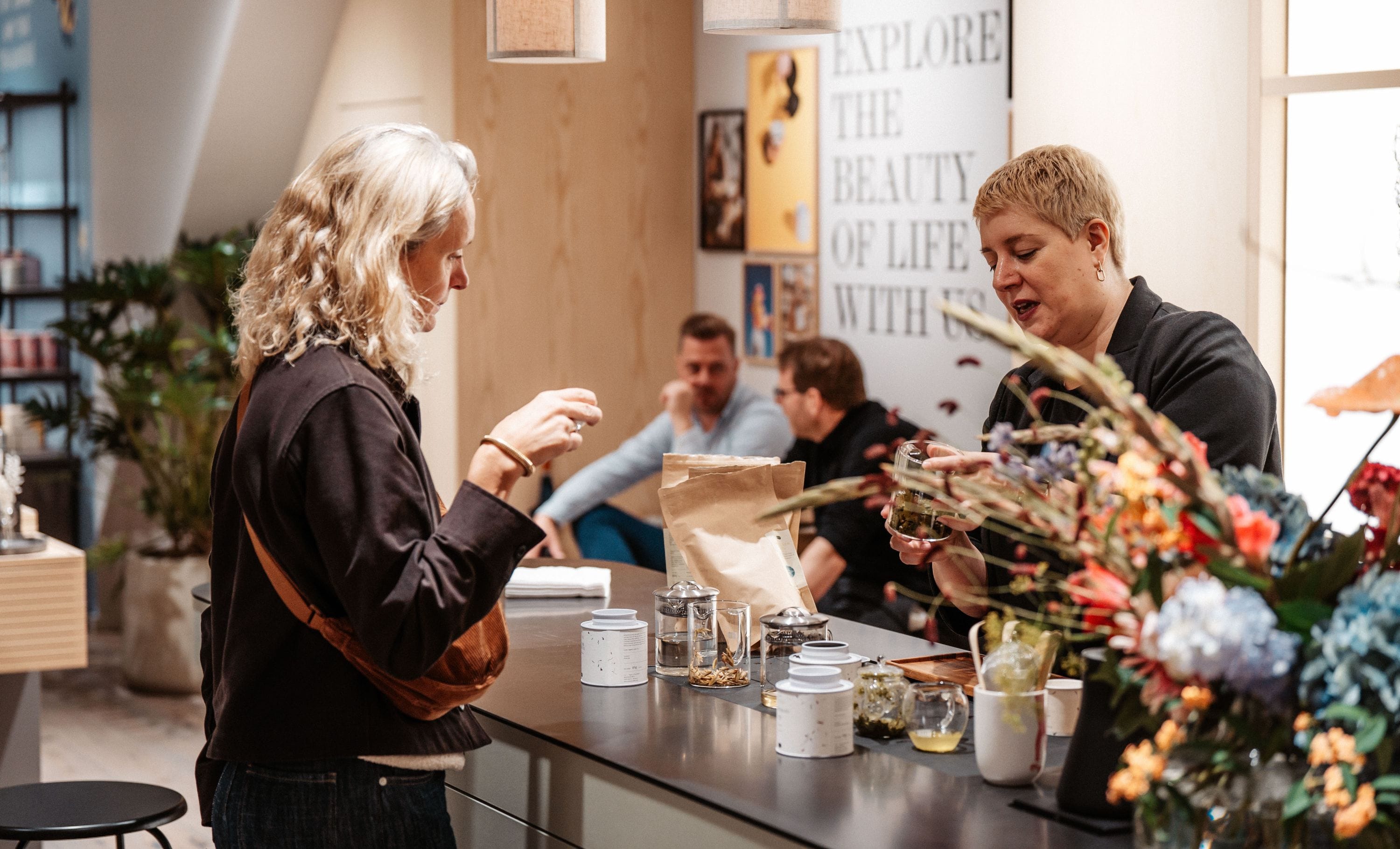 Zwei Frauen an einem Teetisch mit Gläsern und Kräutertees, umgeben von bunten Blumen im Laden.