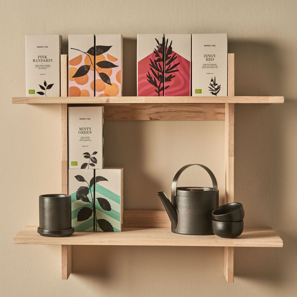 Minimalistisches Holzregal mit Papier- und Teepaketen, schwarzem Keramik-Teeservice mit Teekanne und Tassen.