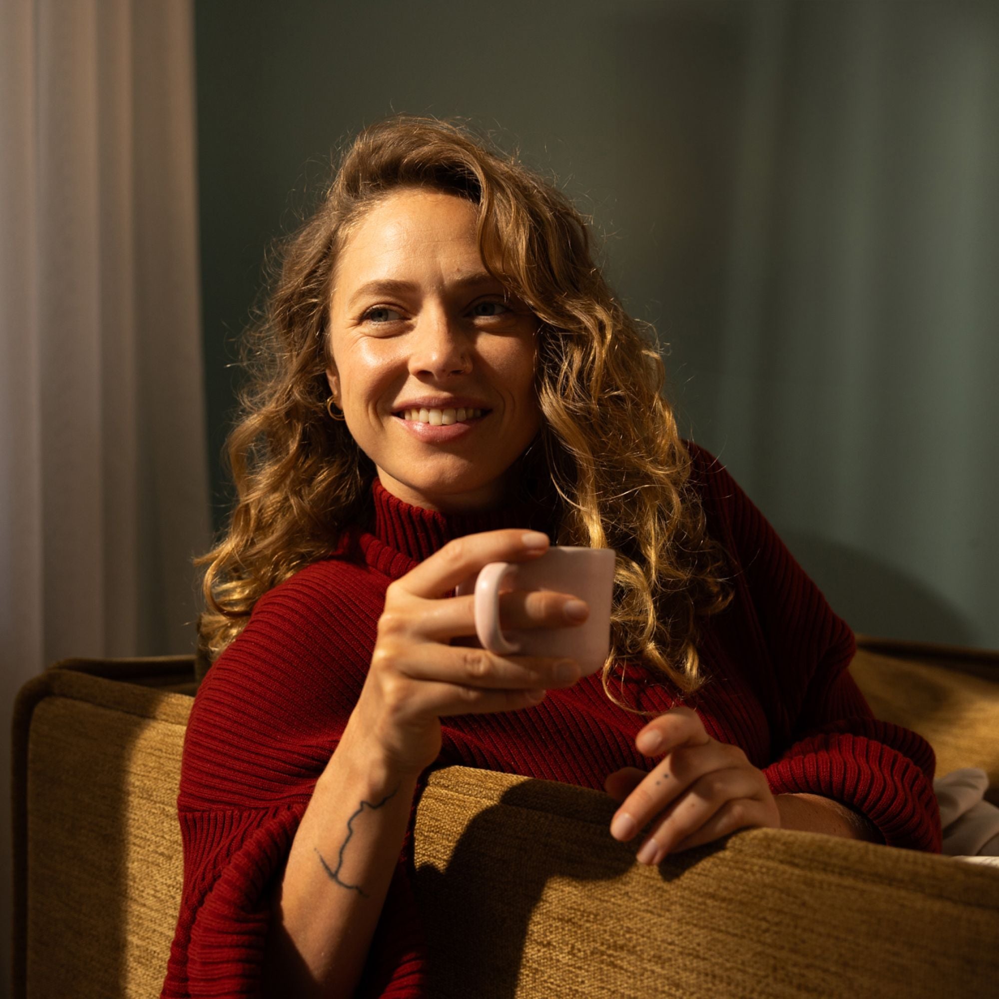 Frau mit lockigen hellbraunen Haaren in rotem Strickpulli, lächelnd mit rosa Becher auf braunem Sofa.