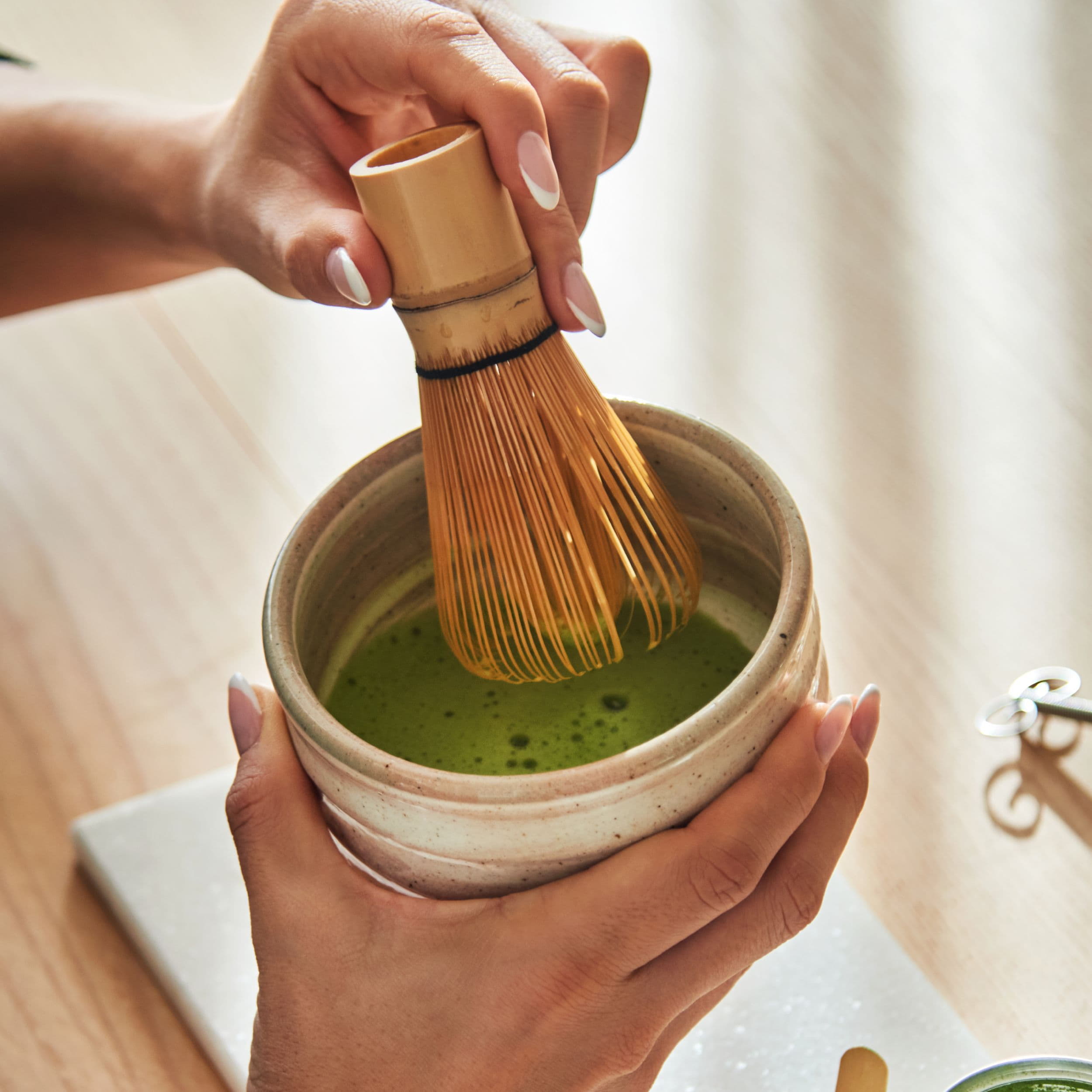 Zwei Hände bereiten cremigen Matcha-Tee in einer Keramikschale mit einem Bambusbesen auf einem Holztisch zu.