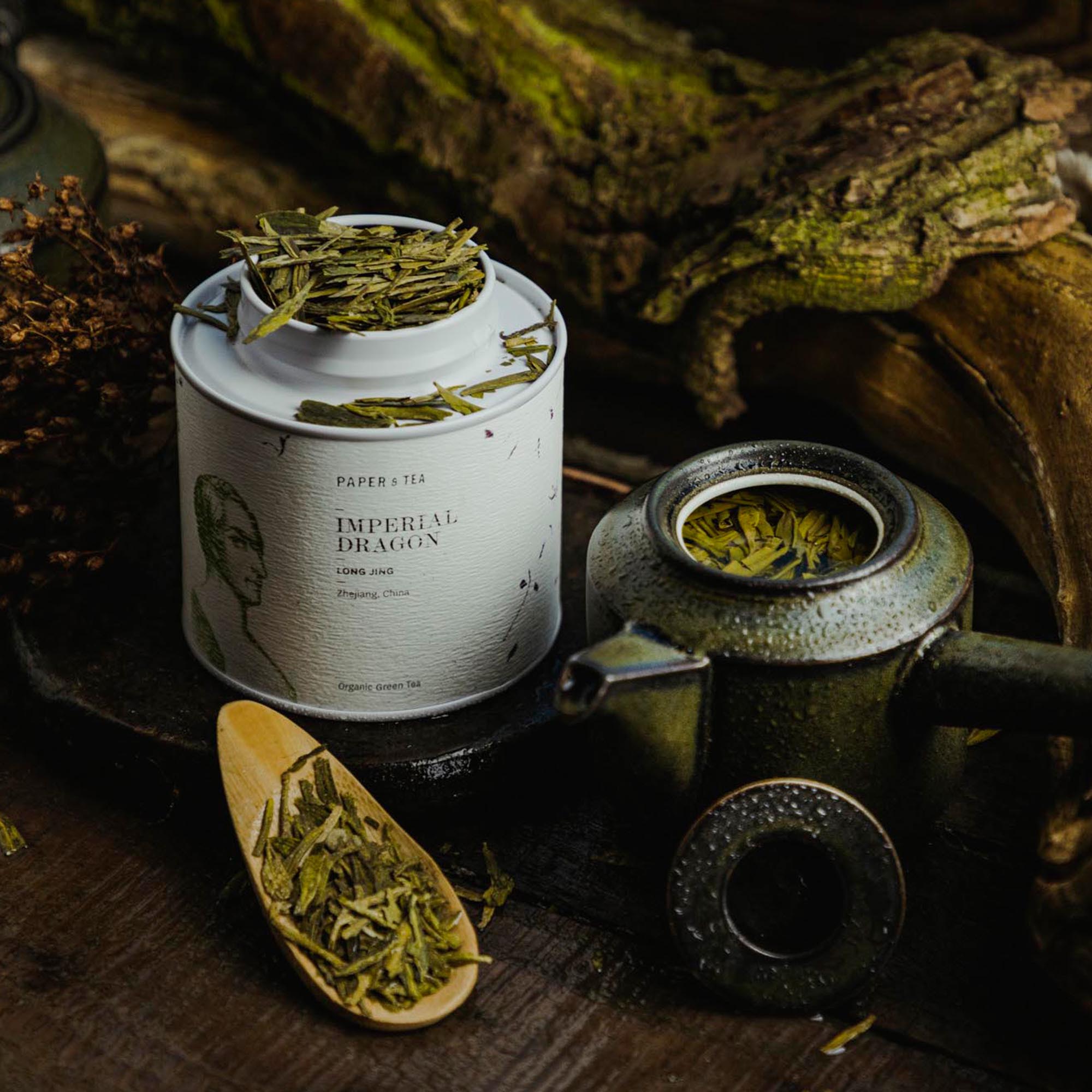 Weiße Teedose mit losem Imperial Dragon Long Jing Bio-Grüntee aus Zhejiang, daneben grüne Keramikkanne und Holzlöffel vor Moos.