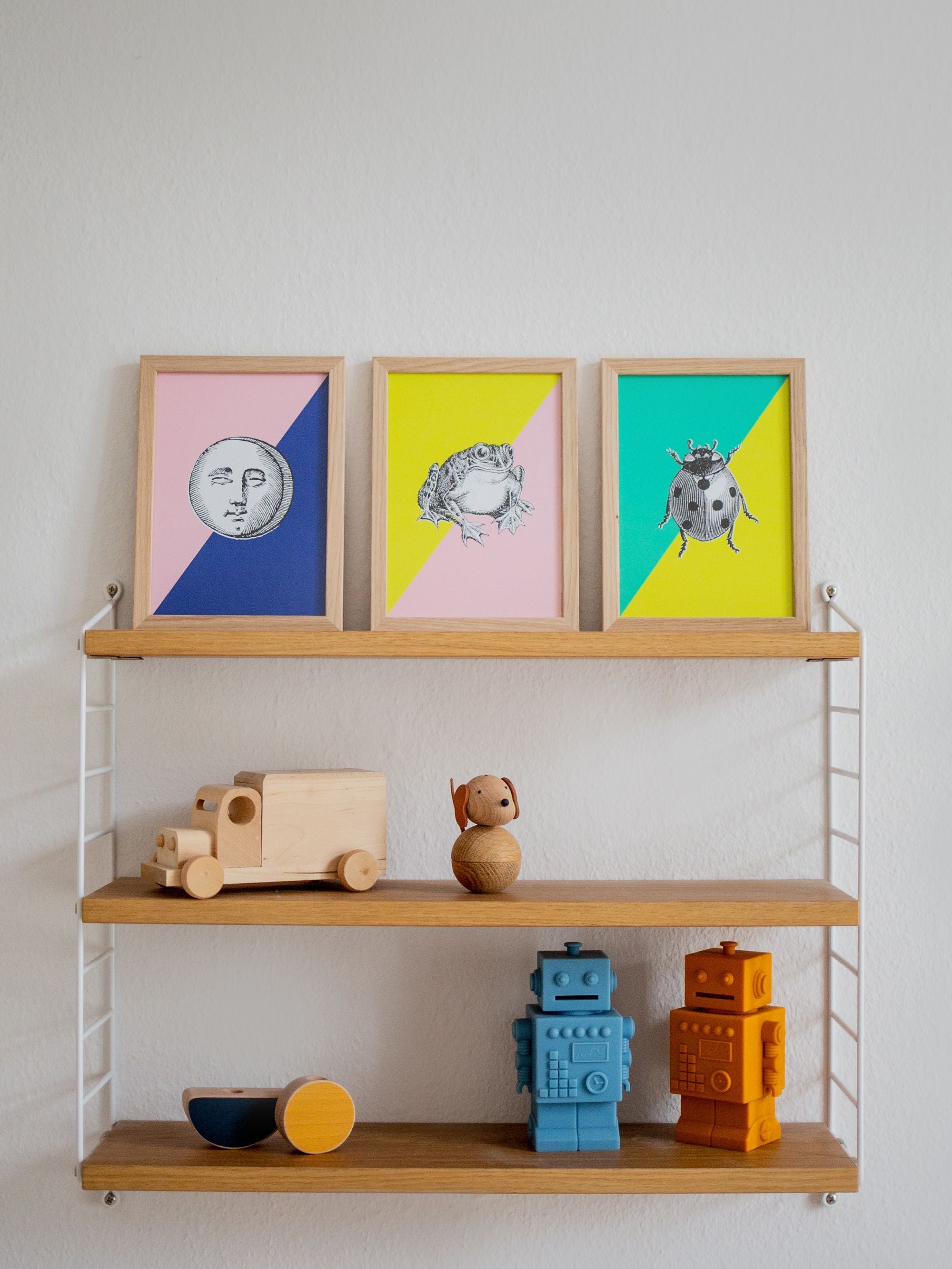 Drei gerahmte Illustrationen: Mondgesicht, Frosch, Marienkäfer auf pastellfarbenen Hintergründen; hölzerne Spielzeuge auf Wandregalen.