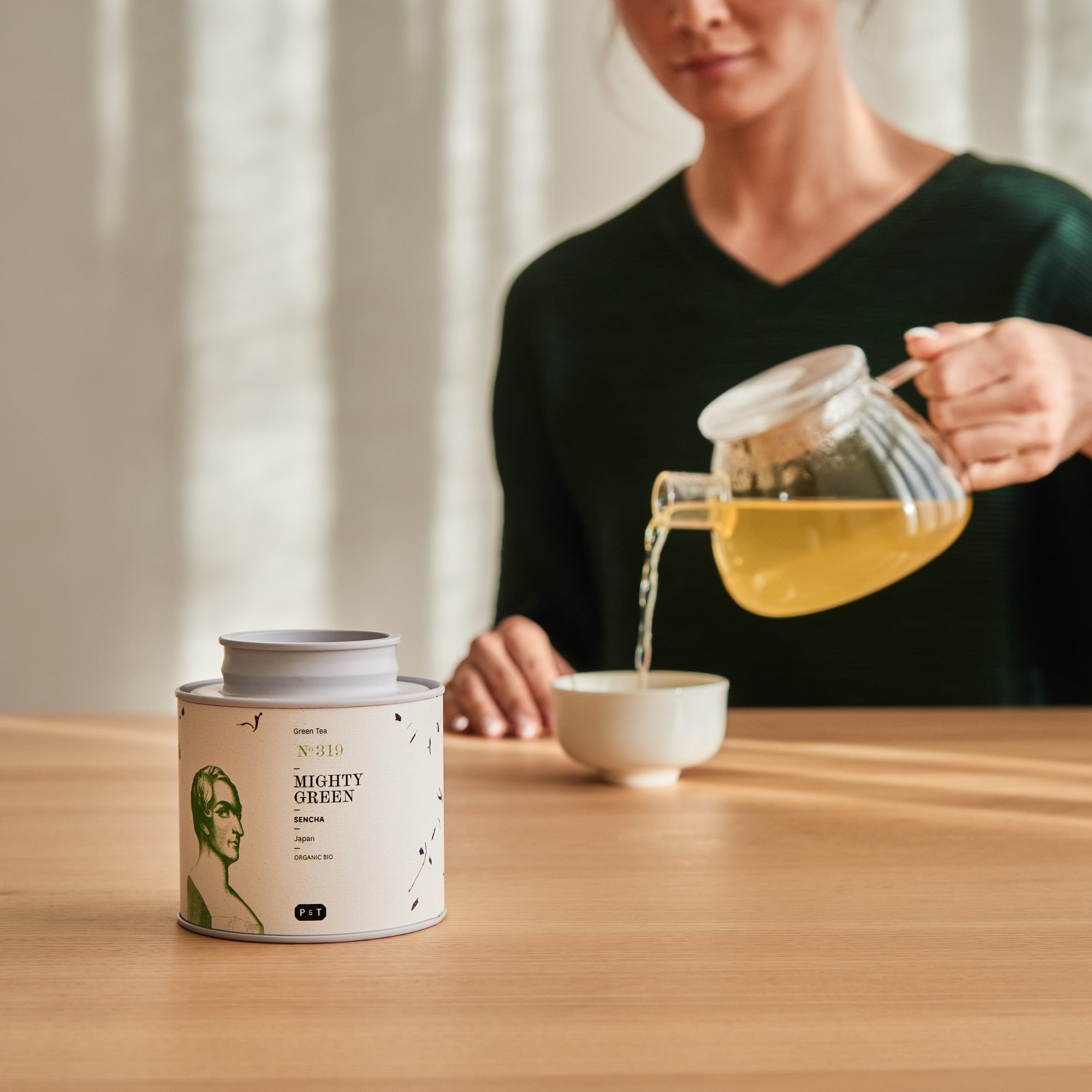 Zylinderförmige Dose mit MIGHTY GREEN Sencha Tee auf Tisch, Person gießt Tee in Tasse im Hintergrund.