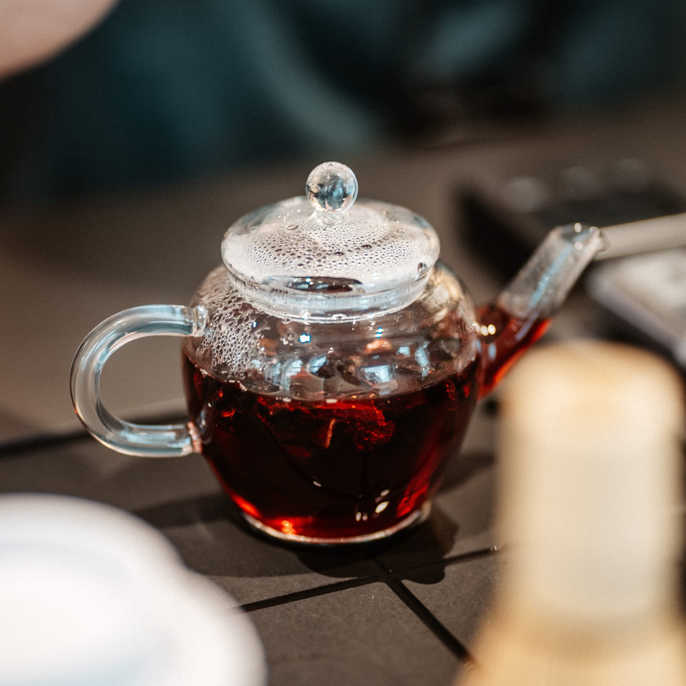 Transparenter Glasteekessel mit dampfendem rotem Tee auf grauer Fliesentischplatte