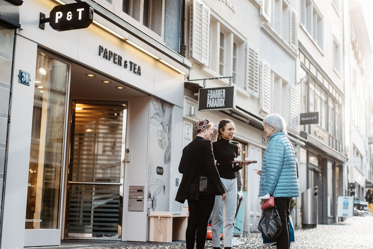 Straßenszene mit dem Teeladen PAPER & TEA, beleuchtetem Eingang und drei Personen im Gespräch.