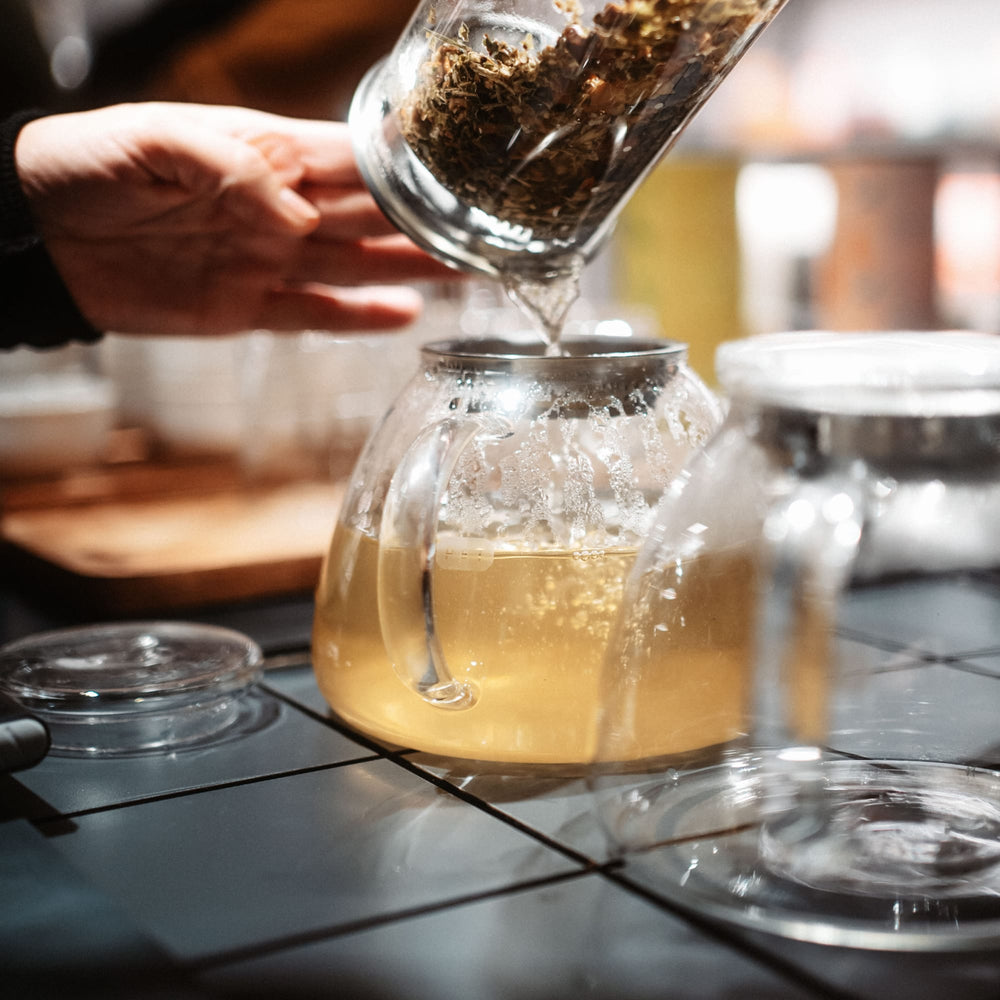 Hand gießt getrocknete Kräuter aus Glasbehälter in Teekanne mit Kräutertee auf Kacheloberfläche.