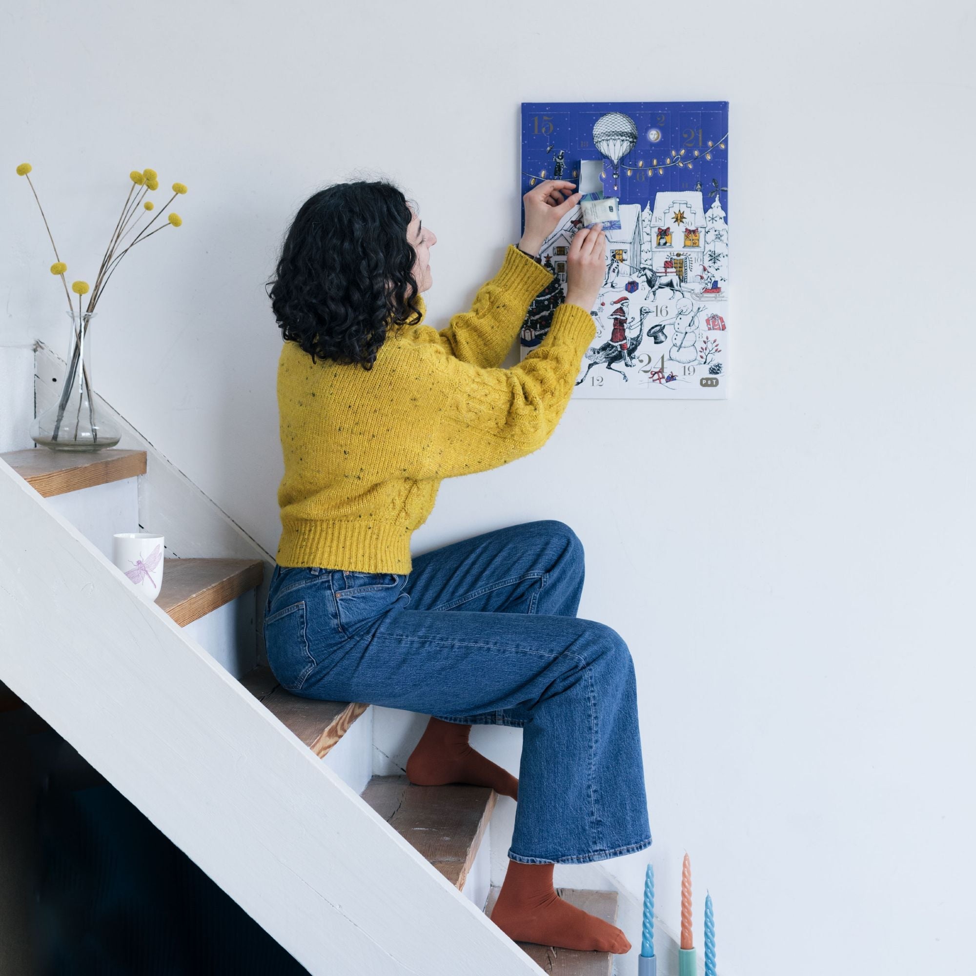 Frau in senfgelbem Pullover öffnet Klappe 16 eines blau-weißen Adventskalenders an weißer Wand.