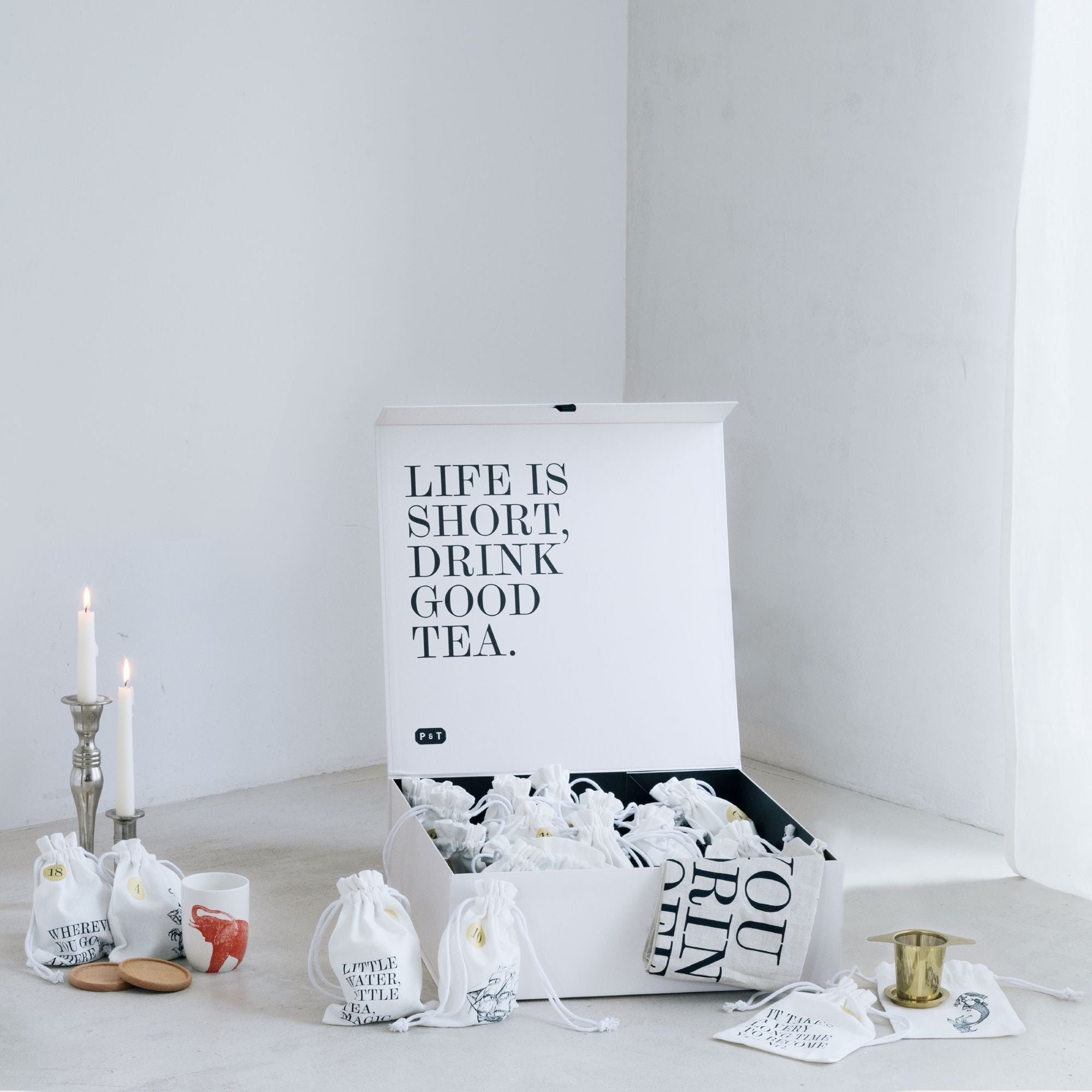 Weiße Teebox mit beschrifteten Baumwollbeuteln, Kerzen, Keramiktasse und goldenem Teesieb