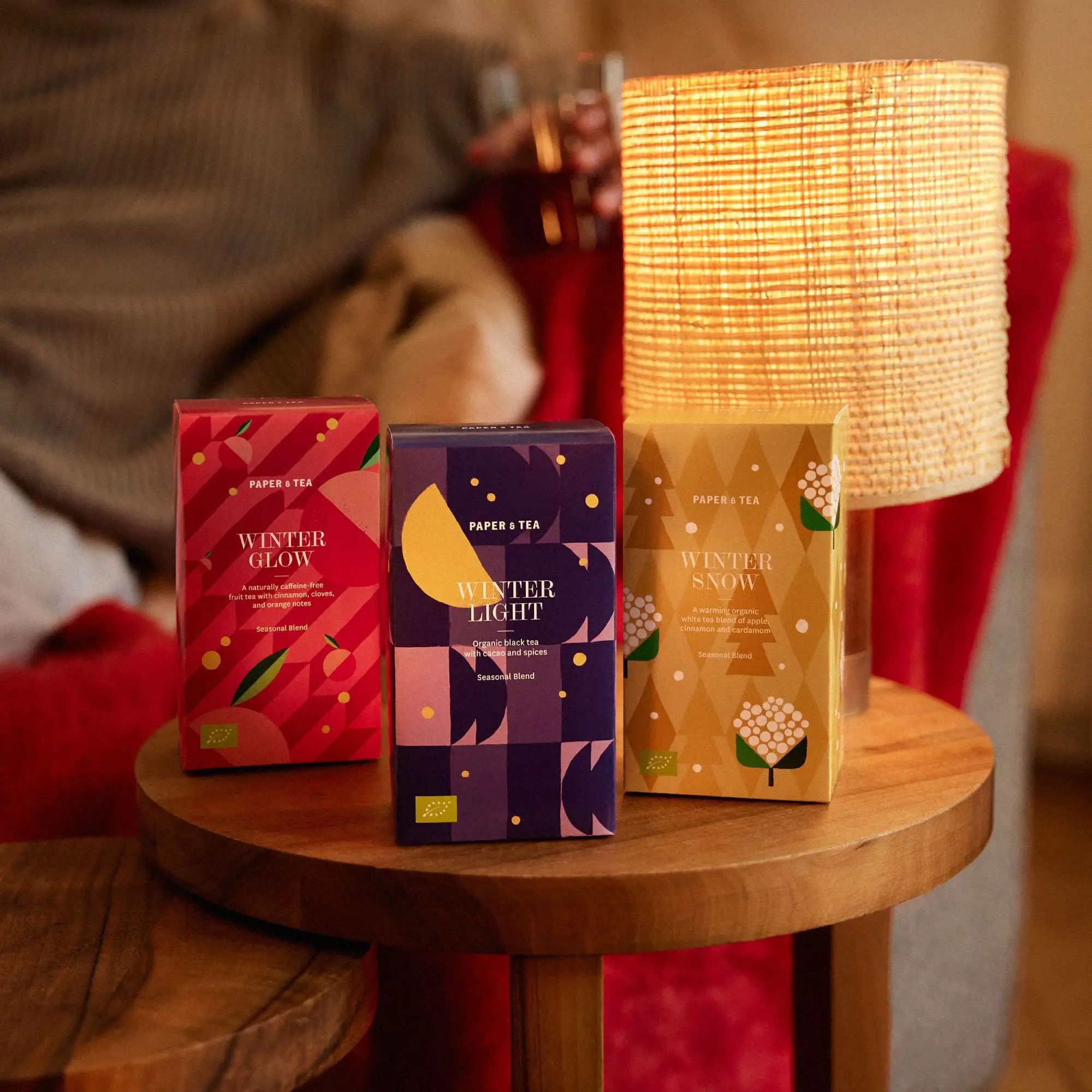 Drei farbenfrohe PAPER & TEA Winter-Teeboxen auf rundem Holztisch mit warmem Licht, verschwommene Person im Hintergrund.