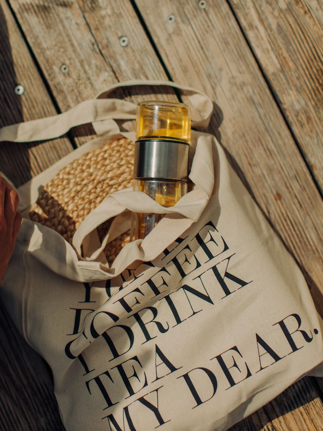 Beige Tragetasche mit schwarzem Aufdruck 'OFFEE DRINK TEA MY DEAR', enthält Flasche mit gelbem Getränk und Strohhalm auf Holzboden.