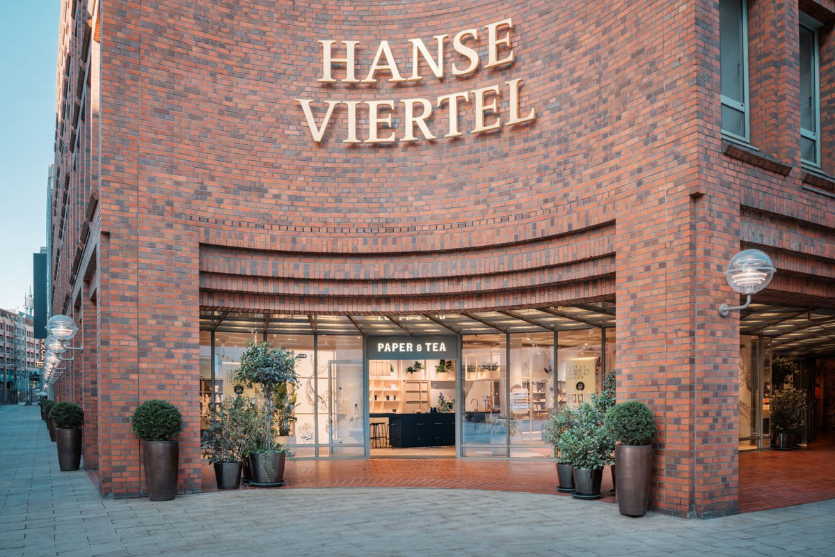 Backsteinfassade mit HANSE VIERTEL Schriftzug, Eingang zu PAPER & TEA, grünen Pflanzen und Beleuchtung.