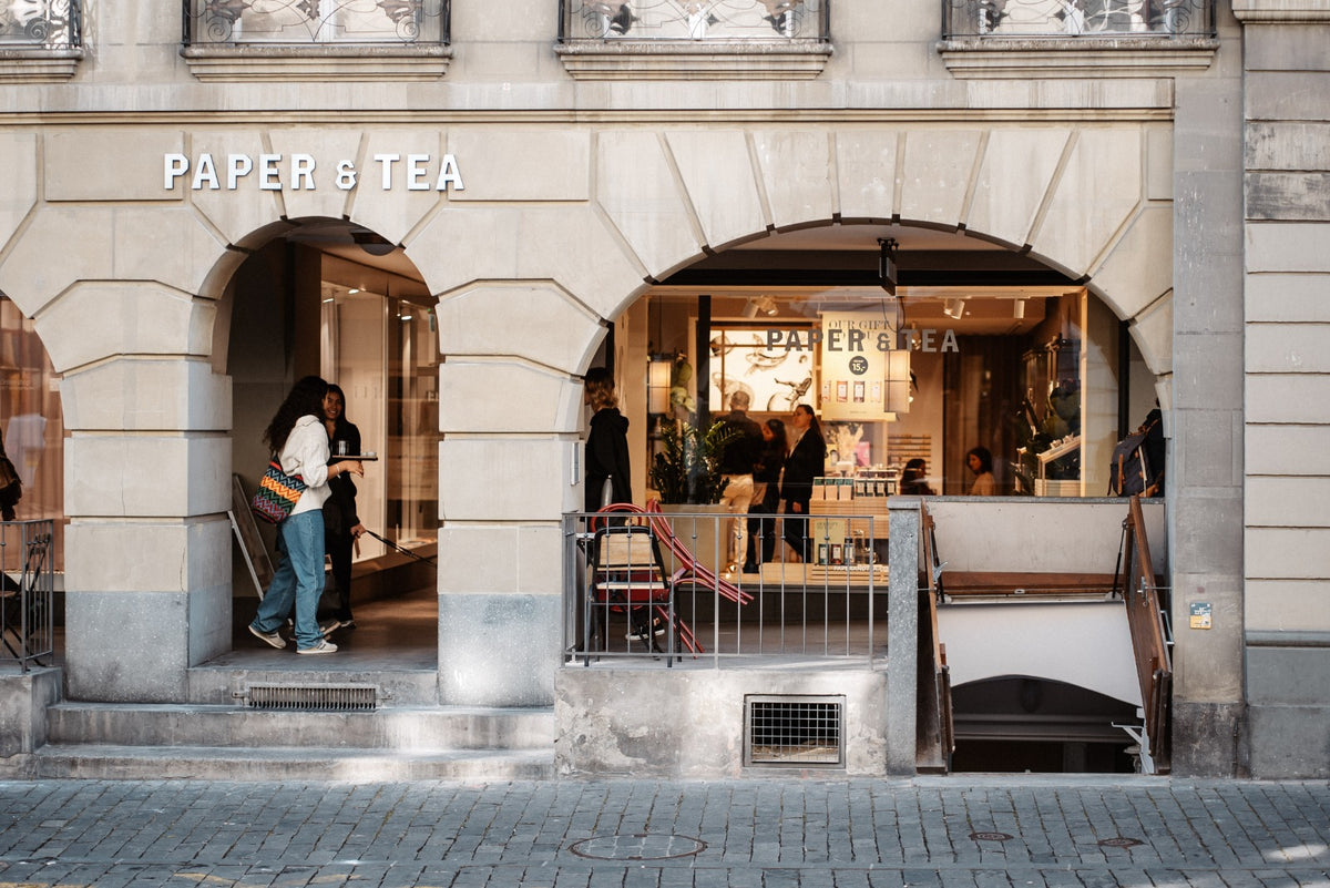 Eingang des PAPER & TEA Ladens mit Steinbogenfassade, Kunden davor und historischem Kopfsteinpflaster.