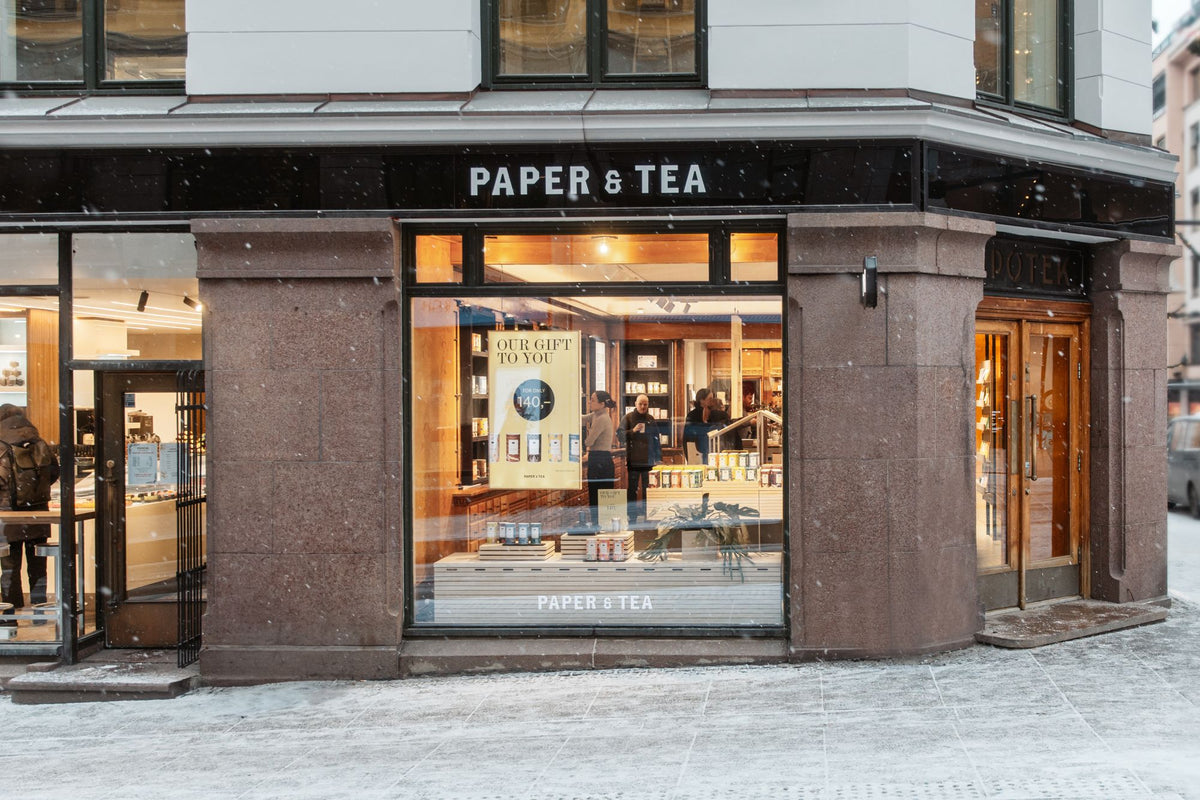 Hell erleuchteter PAPER & TEA Laden mit großen Schaufenstern, gefüllt mit hochwertigen Tees; Schneeszene davor.